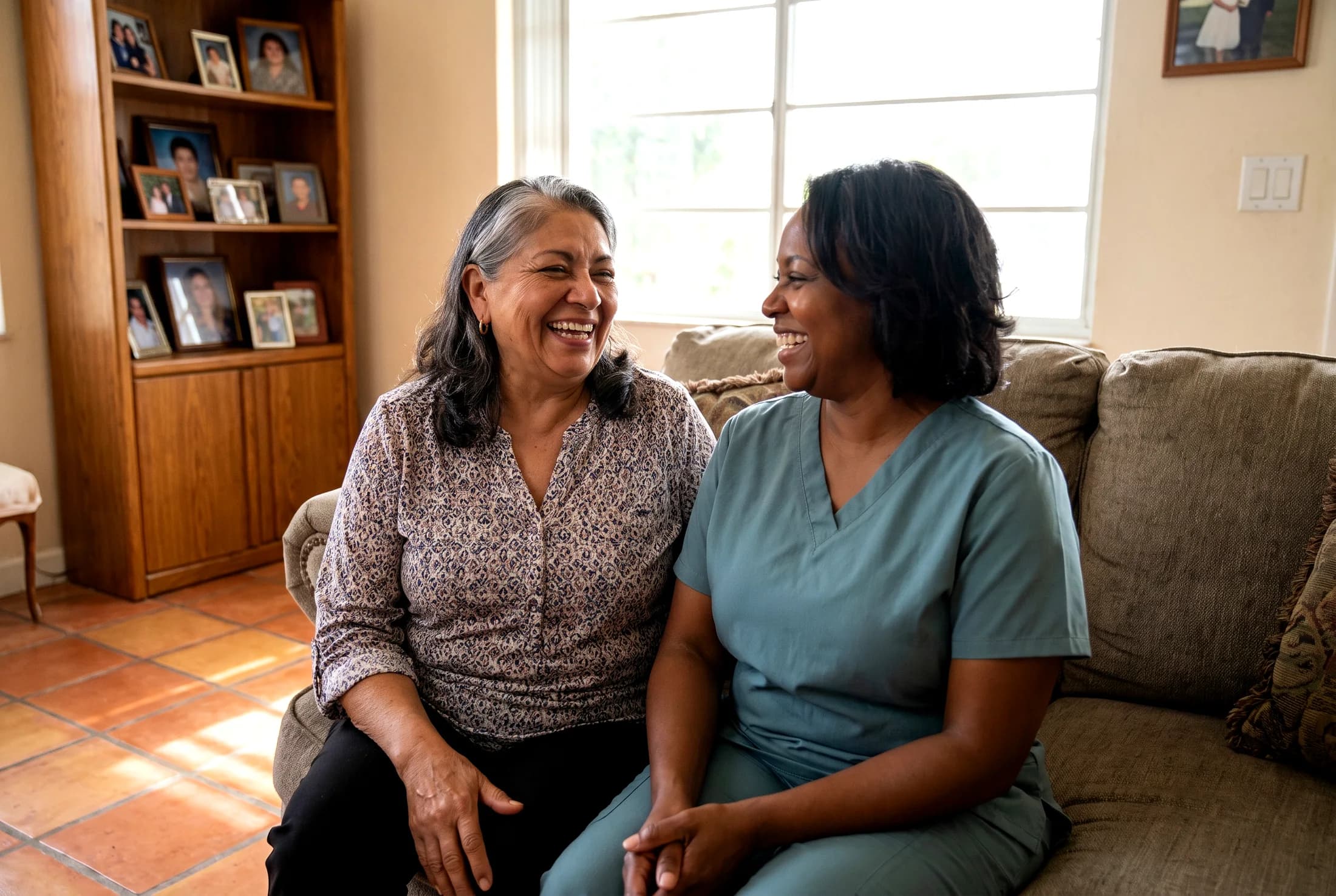 A caregiver in a teal uniform sitting with an older woman on a sofa, both laughing
