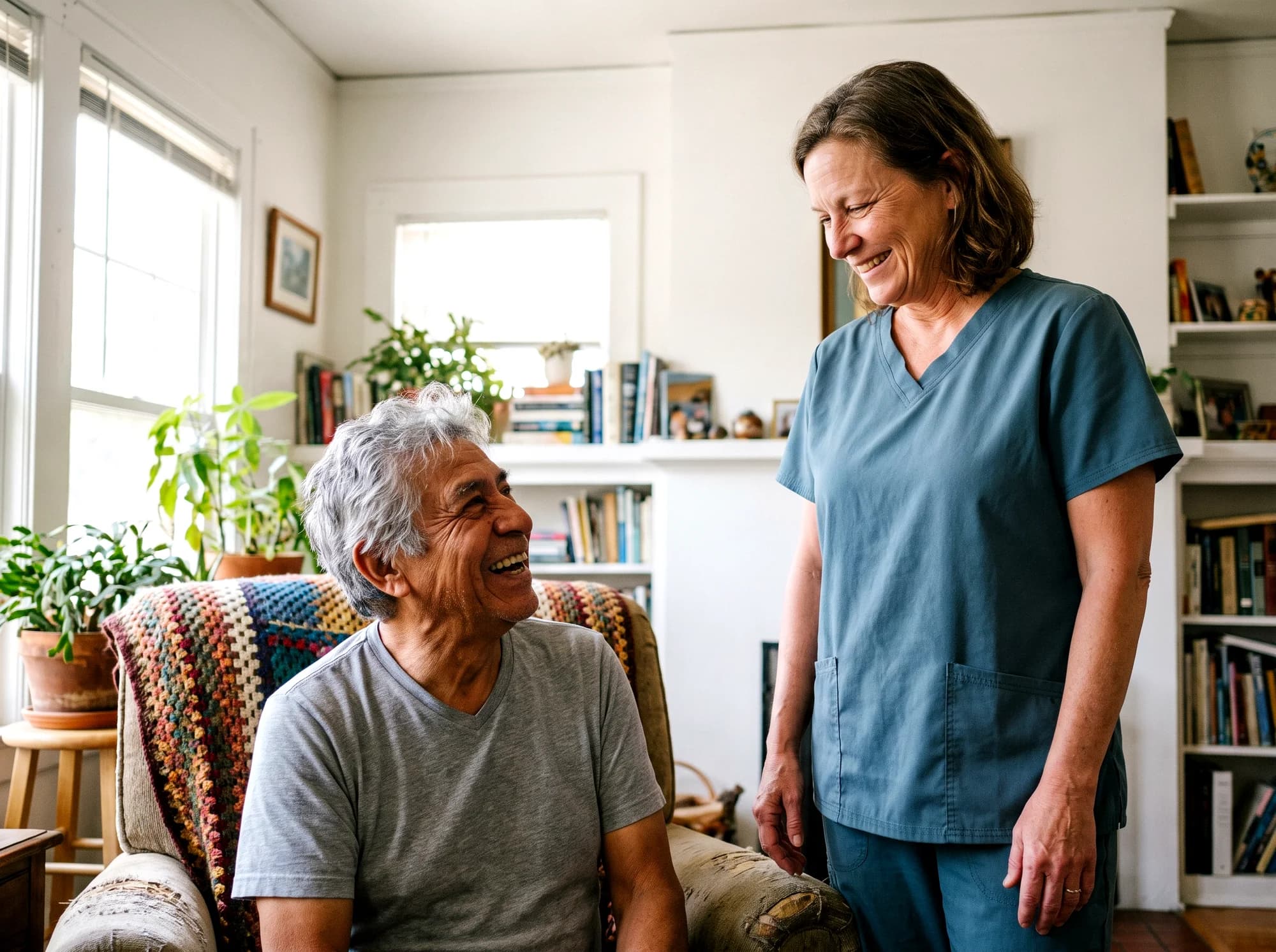 A caregiver in a teal uniform standing beside an older man in an armchair, both smiling