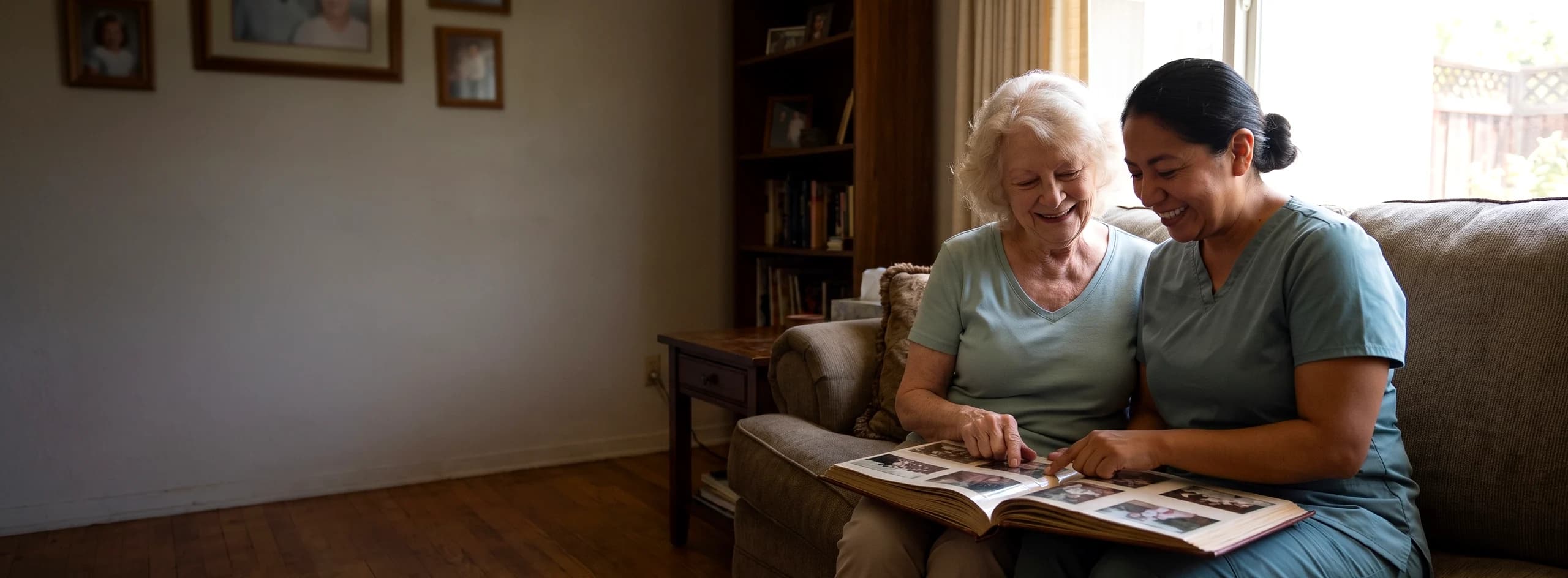 A caregiver in teal scrubs and an older woman sitting together on a sofa at home, smiling over an open photo album
